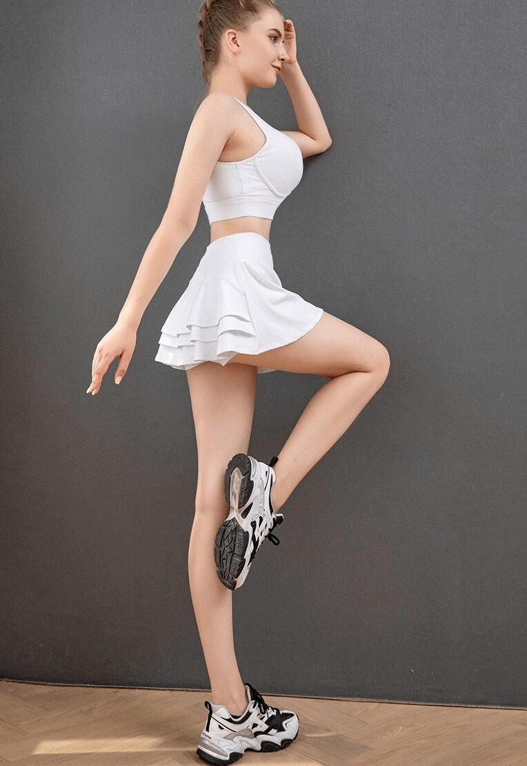 Woman wearing a white pleated tennis skirt with inside pocket, demonstrating flexibility, against a gray background.